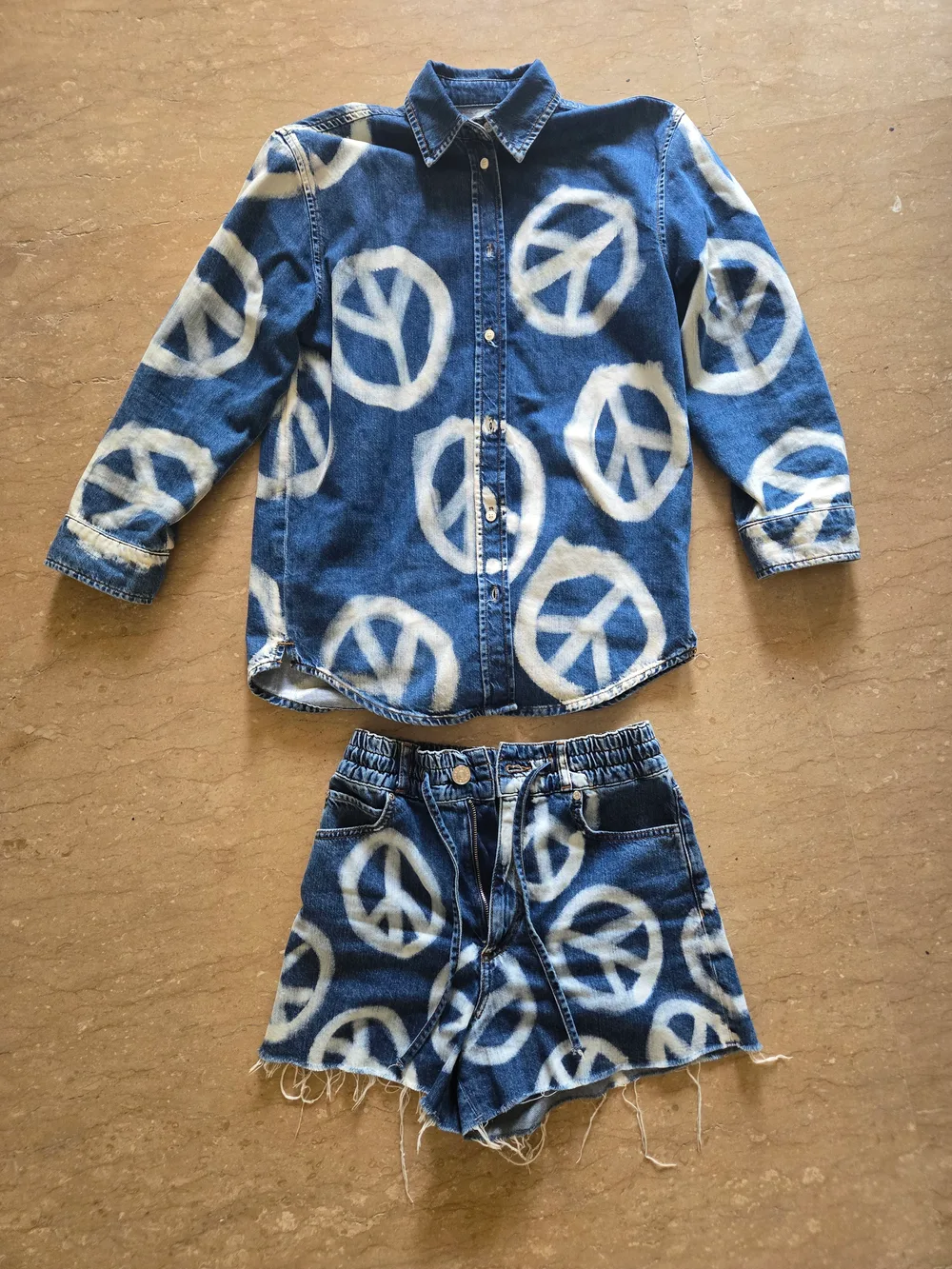 Input flat lay, front. A bleached peace print denim shirt above matching cut off shorts on a tan floor, showing the button placket, the shirred waistband, the drawstring, the shank button, the open zip fly and the frayed hem