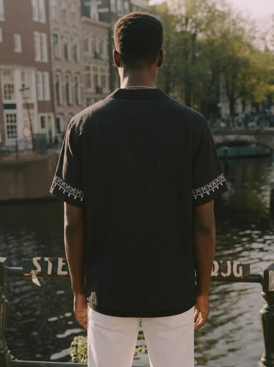 MODA AI output shot 4 of 10 from the same one-credit generation on the black embroidered shirt  — brand model matching uploaded face reference, Amsterdam canal scene matching uploaded background reference