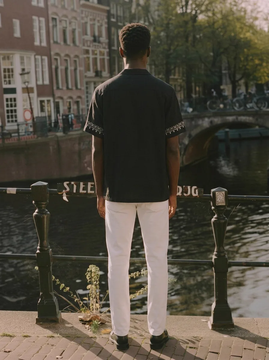 MODA AI output shot 8 of 10 from the same one-credit generation on the black embroidered shirt — brand model matching uploaded face reference, Amsterdam canal scene matching uploaded background reference